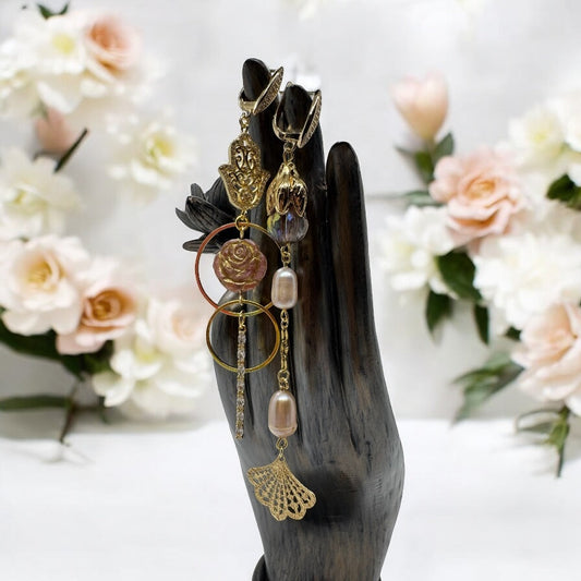 A statue of a hand holding up a pair of mismatched earrings. The earrings have a focal rose bead, a hamsa hand, and pearls.