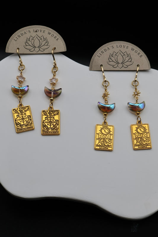 A pair of gold-toned dangle earrings displayed on a white stand against a black background. Each earring has a small faceted bead, a crescent moon charm, and a rectangular engraved plate with the words 'LOVE WORK' and a lotus symbol.