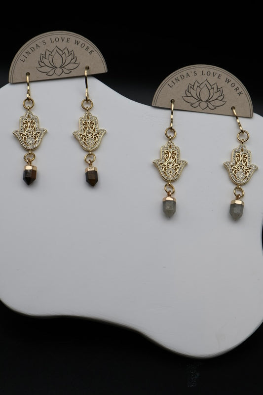 A pair of gold Hamsa hand dangle earrings with dark brown and gray bead drops, displayed on a white stand with brand card inserts.