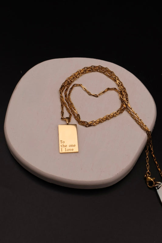 A gold chain necklace with a rectangular gold pendant engraved with 'To the one I love', displayed on a light gray stone plate against a black background.