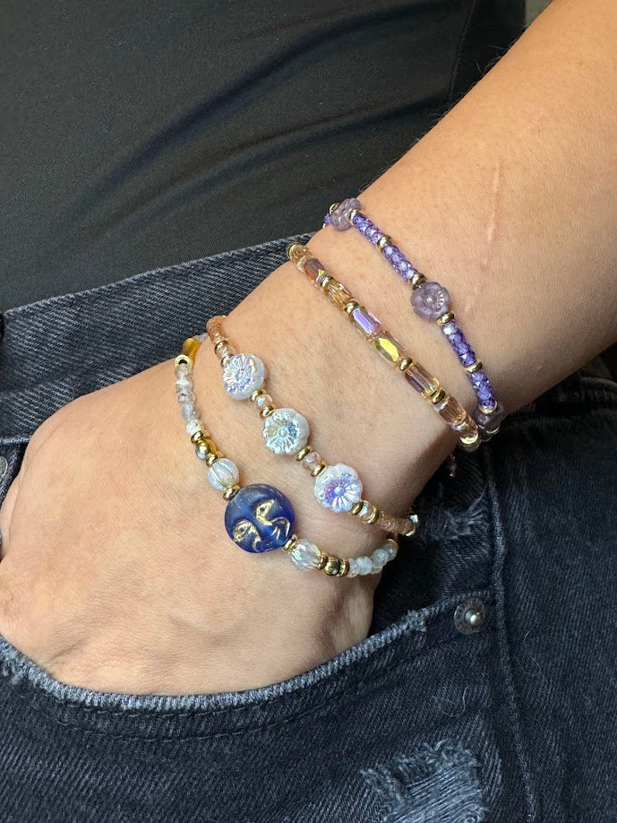 Four bracelets on a wrist. One with a moon face, flowers, peach colored stones, and purple with flowers.