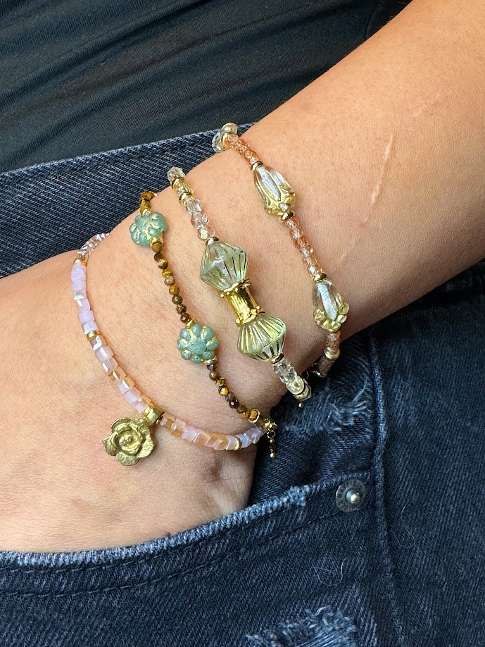 A wrist wearing a bracelet with clear beads and baroque center beads, a bracelet with peach stones and clear tulip beads, a bracelet with tigers eye and green flowers, pink bracelet with a gold rose charm.