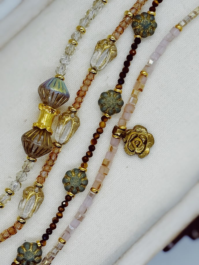 A close up of a bracelet with clear beads and baroque center beads, a bracelet with peach stones and clear tulip beads, a bracelet with tigers eye and green flowers, pink bracelet with a gold rose charm.