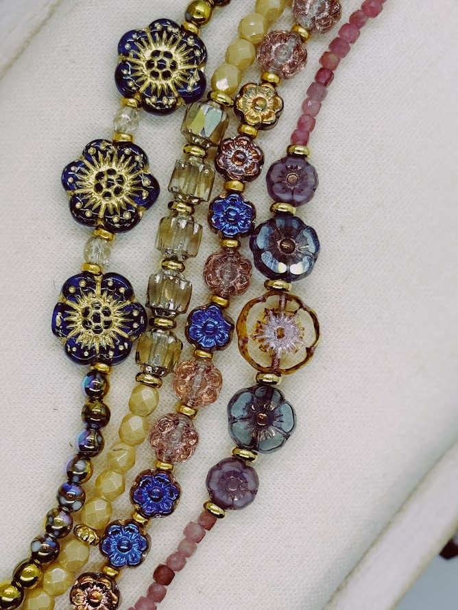 A closeup of a bracelet with three blue flowers and iridescent beads, bracelet with nude beads and baroque center beads, bracelet with metallic flowers, a bracelet with pink stones and five flowers in the center