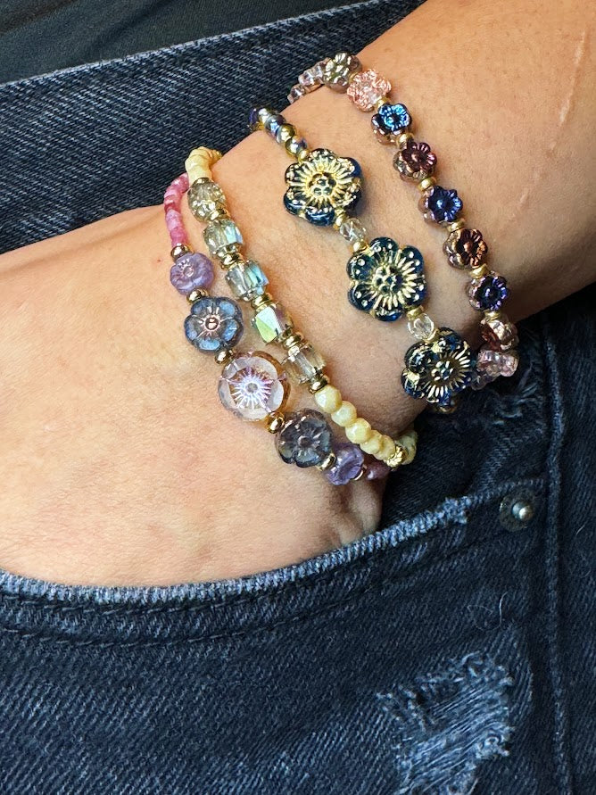 A wrist wearing a bracelet with three blue flowers and iridescent beads, bracelet with nude beads and baroque center beads, bracelet with metallic flowers, a bracelet with pink stones and five flowers in the center