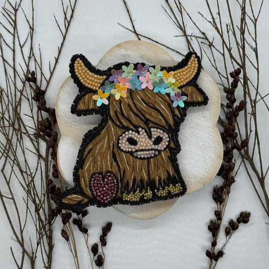 An embroidered pin of a highland cow with a crown of flowers