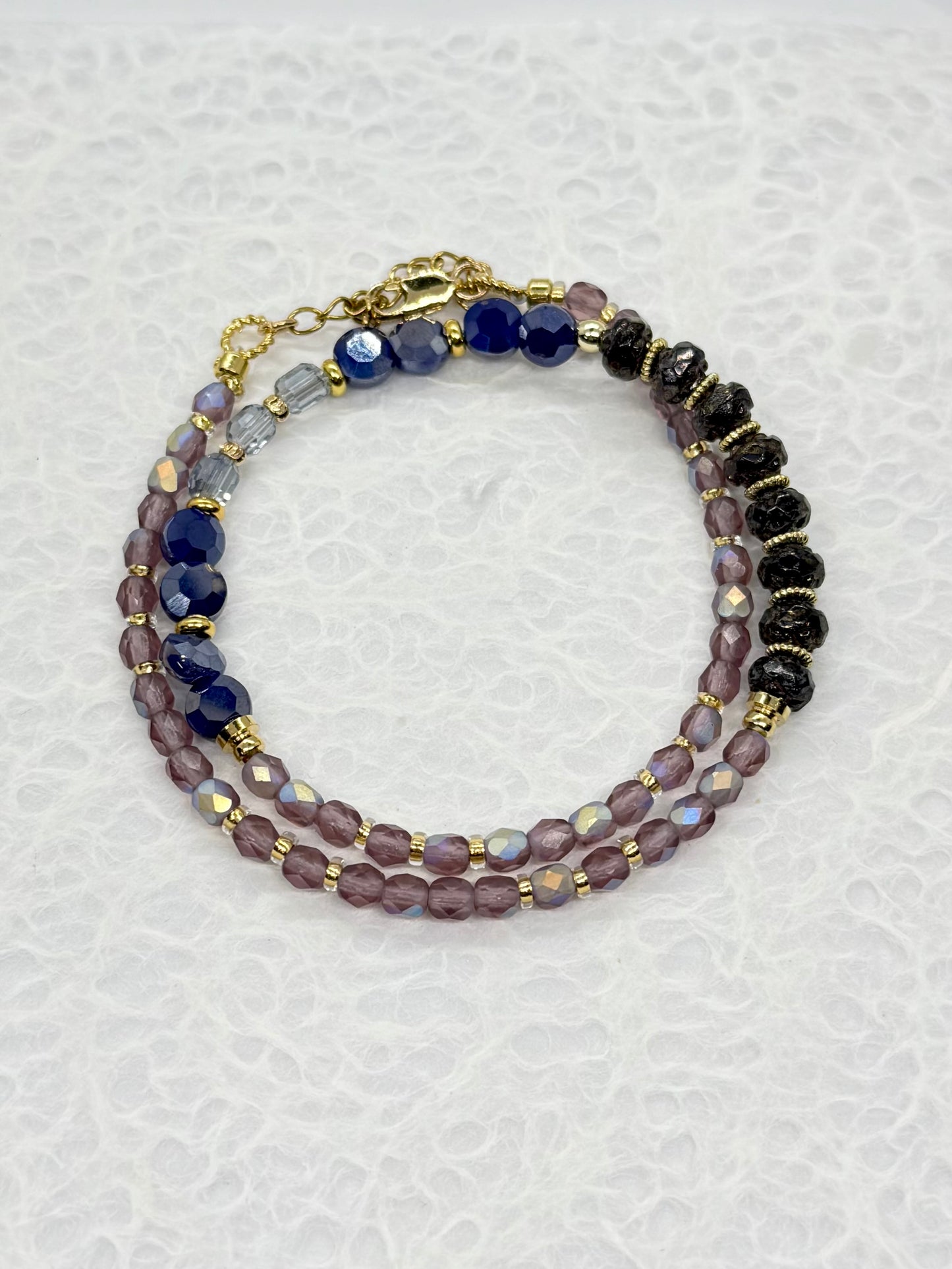 Multistrand beaded bracelet with purple, black, and gold beads on a textured white background