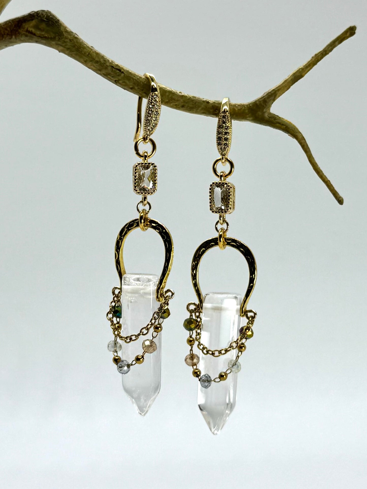 Gold and clear crystal earrings hanging from a branch against a light background