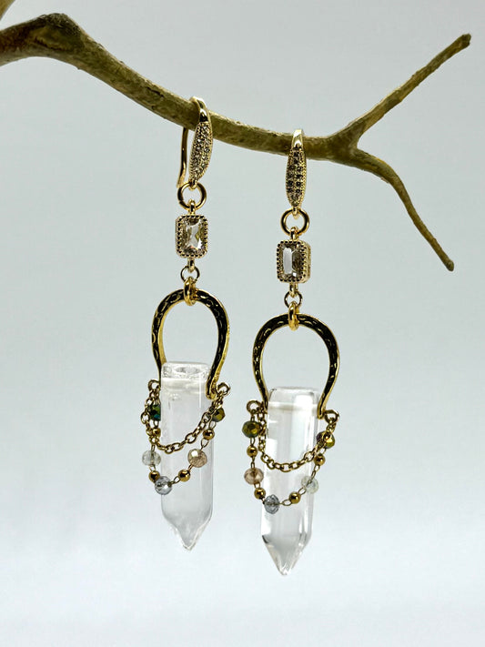 Gold and clear crystal earrings hanging from a branch against a light background