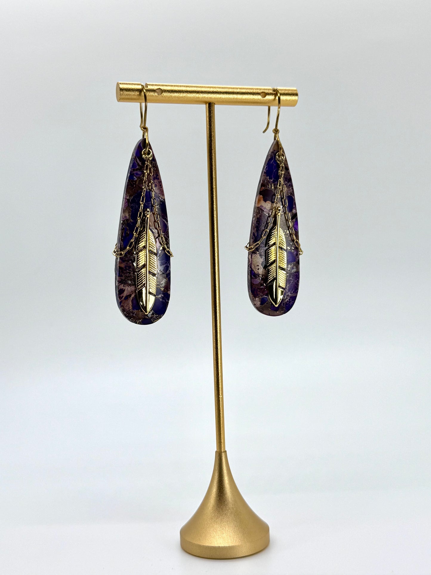 Teardrop-shaped earrings with gold feather design on a brass stand against a white background