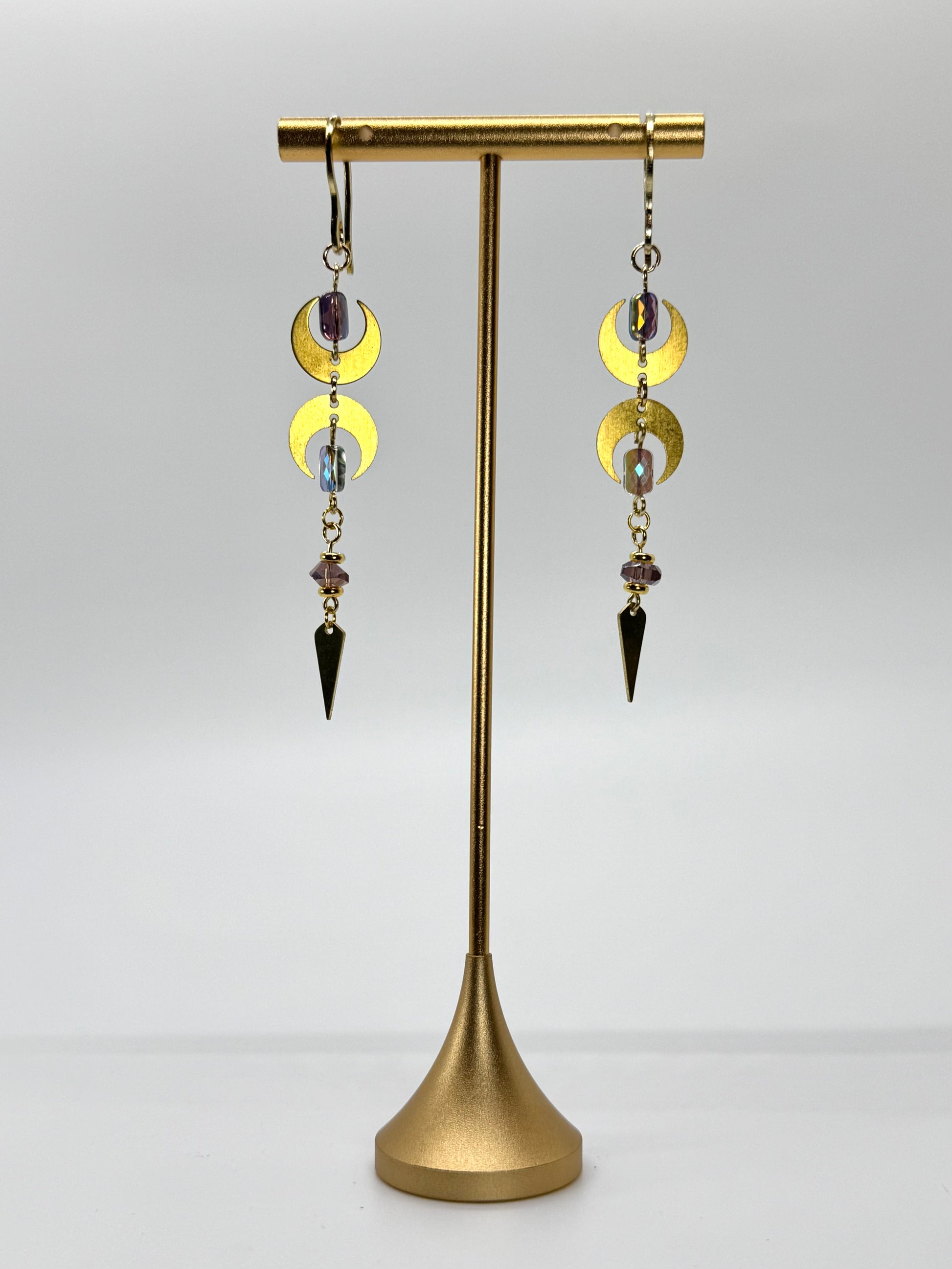 Pair of gold earrings with circular and triangular charms on a gold stand against a white background