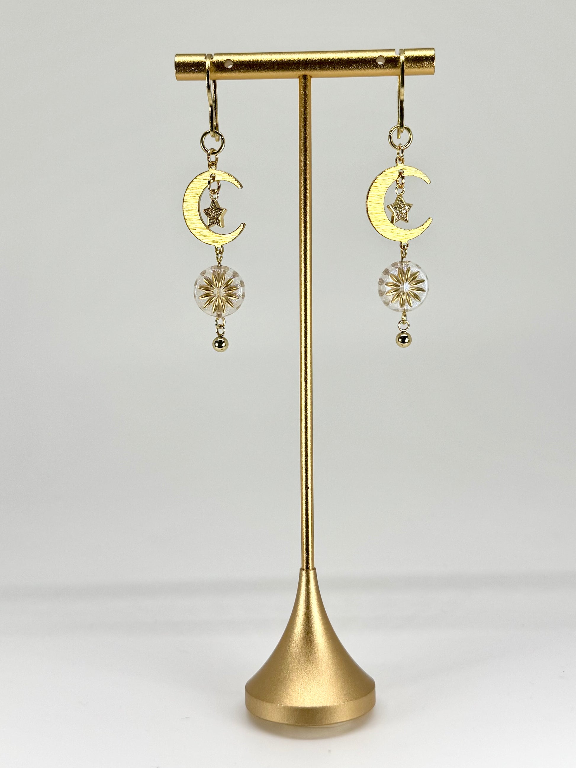 Gold earrings with moon and star designs on a gold stand against a white background