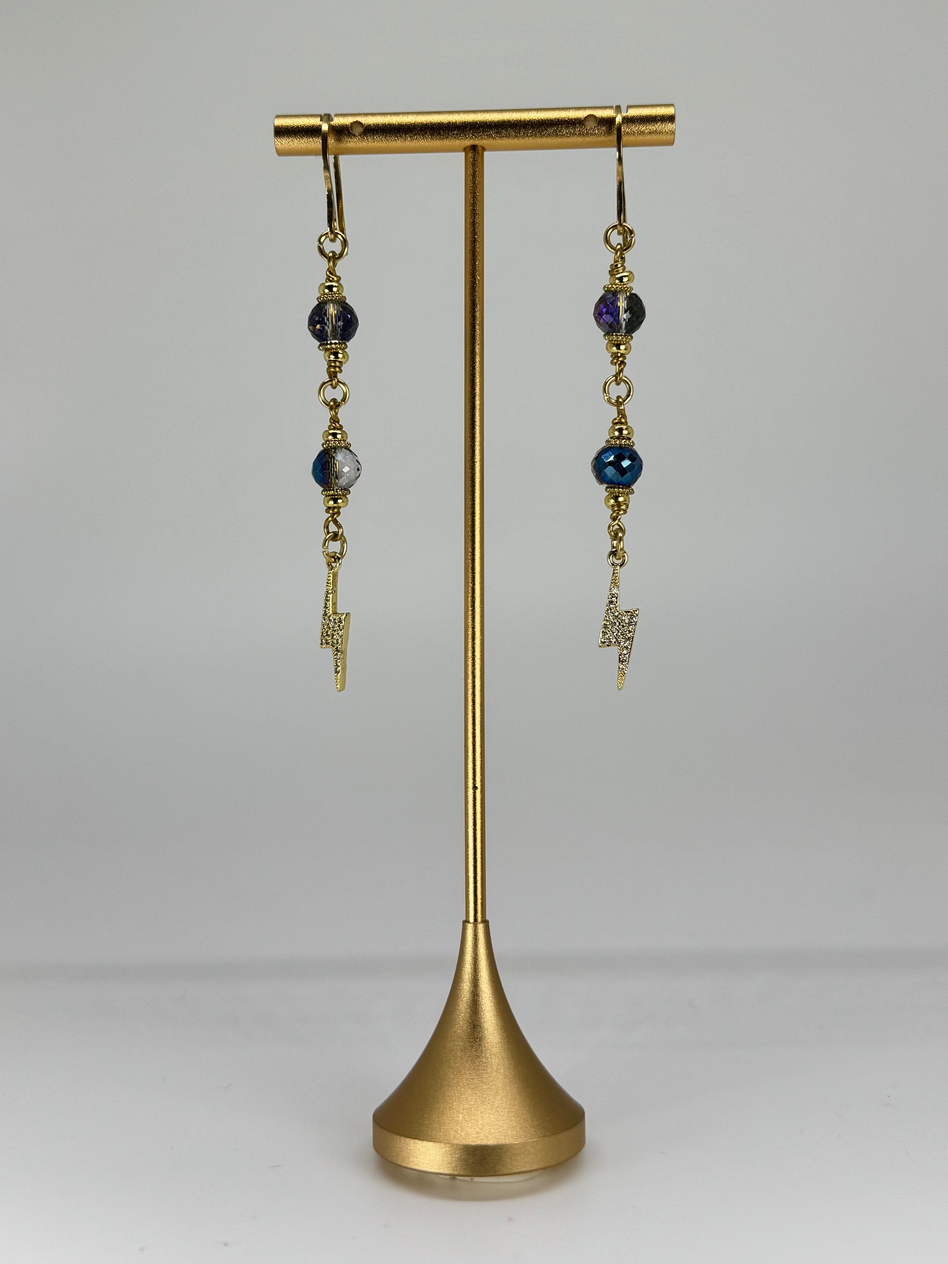 Pair of earrings with blue beads and gold accents on a brass stand against a gray background