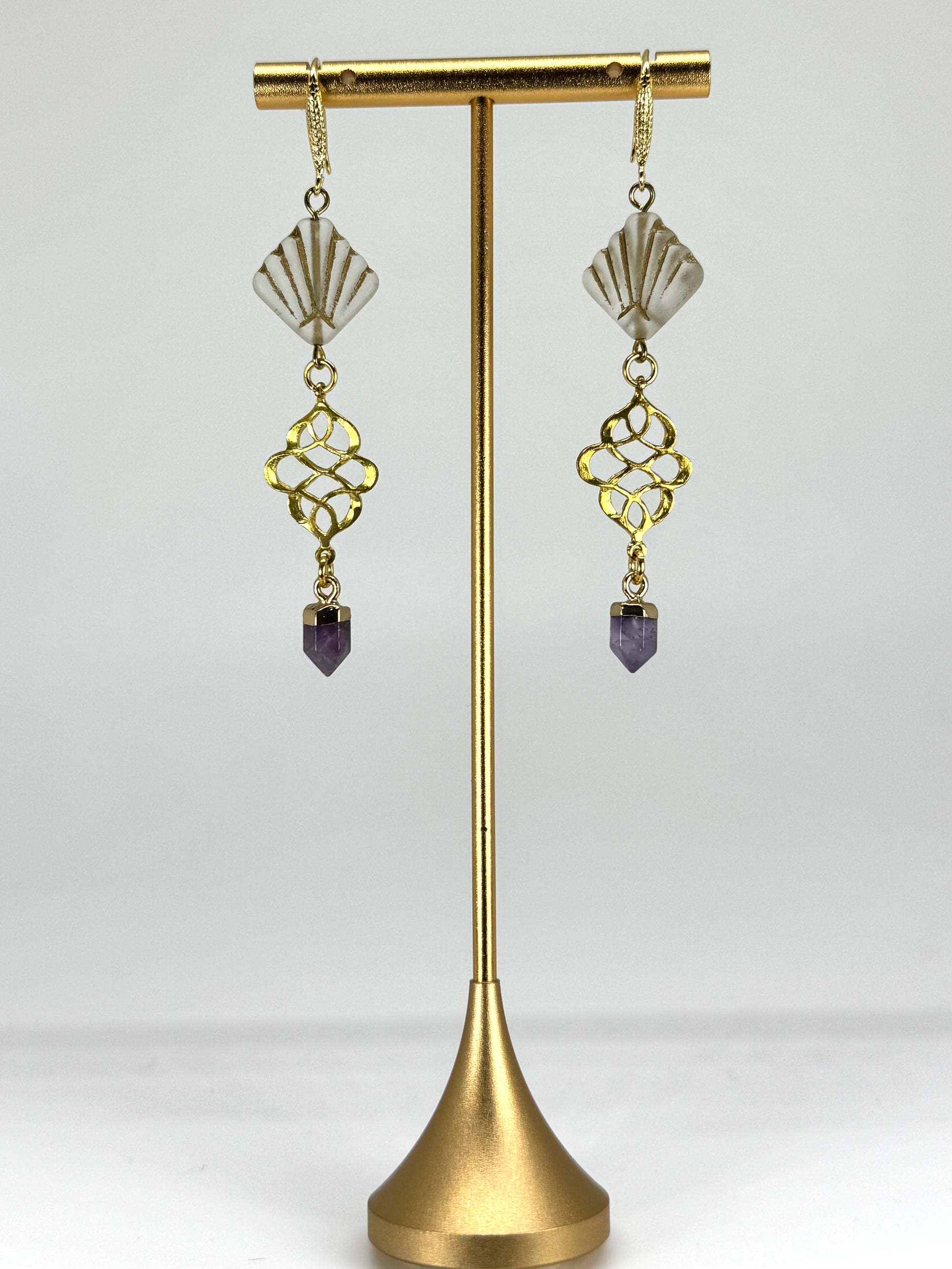 Gold earrings with purple stones on a gold stand against a white background