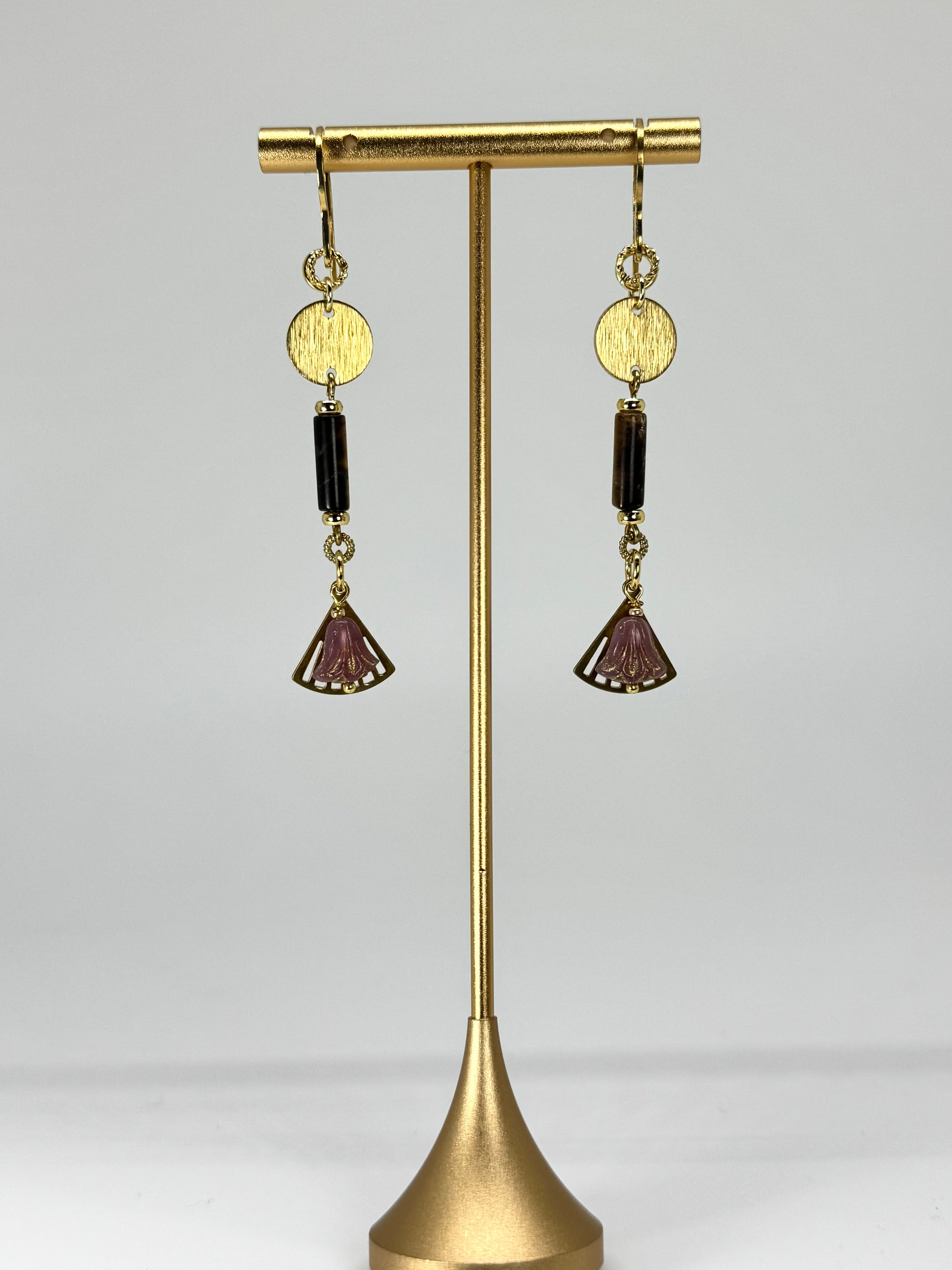 Gold earrings with purple and black stones on a gold stand against a light gray background