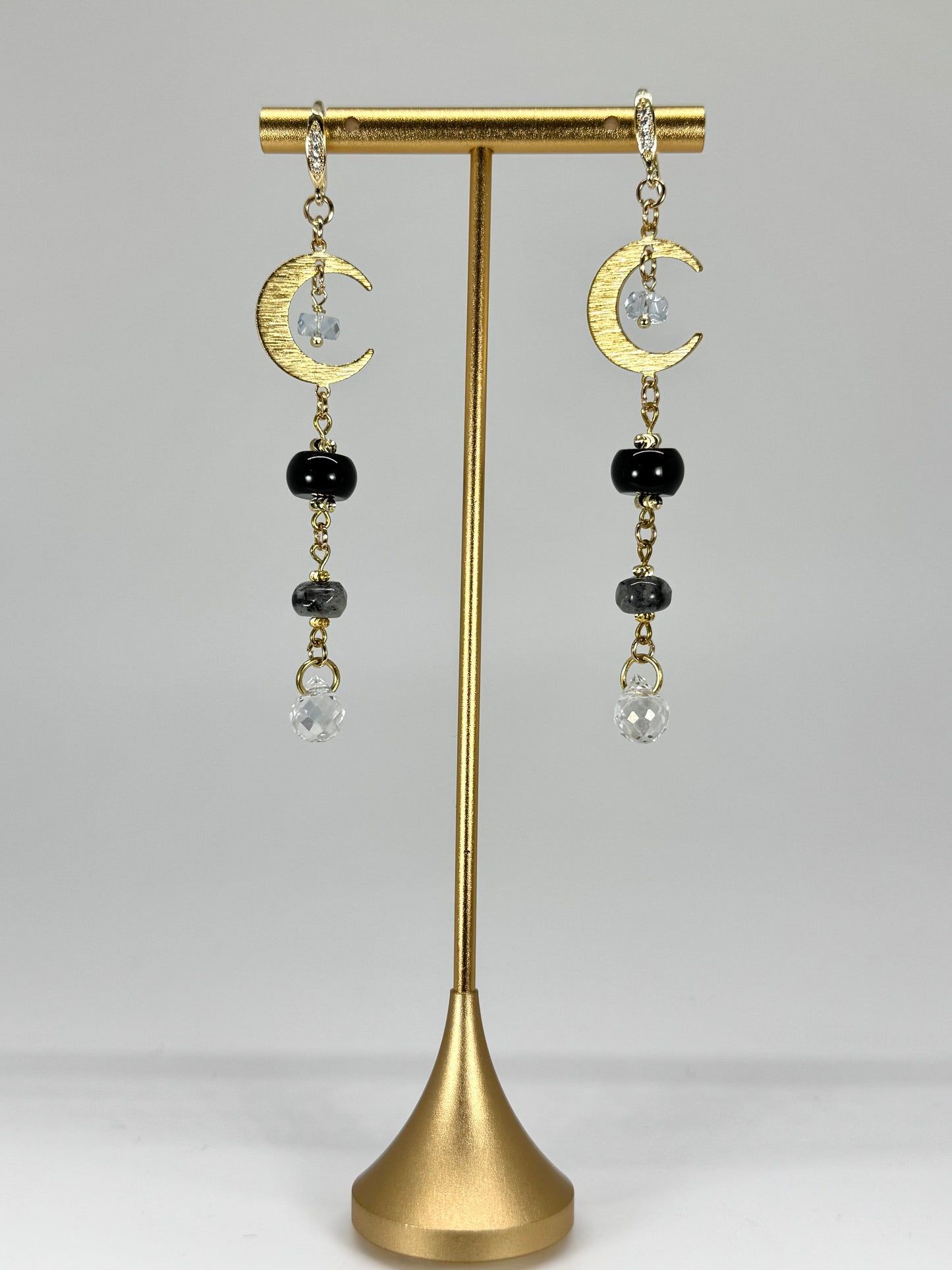 Gold earrings with crescent moon and black bead designs on a gold stand against a gray background