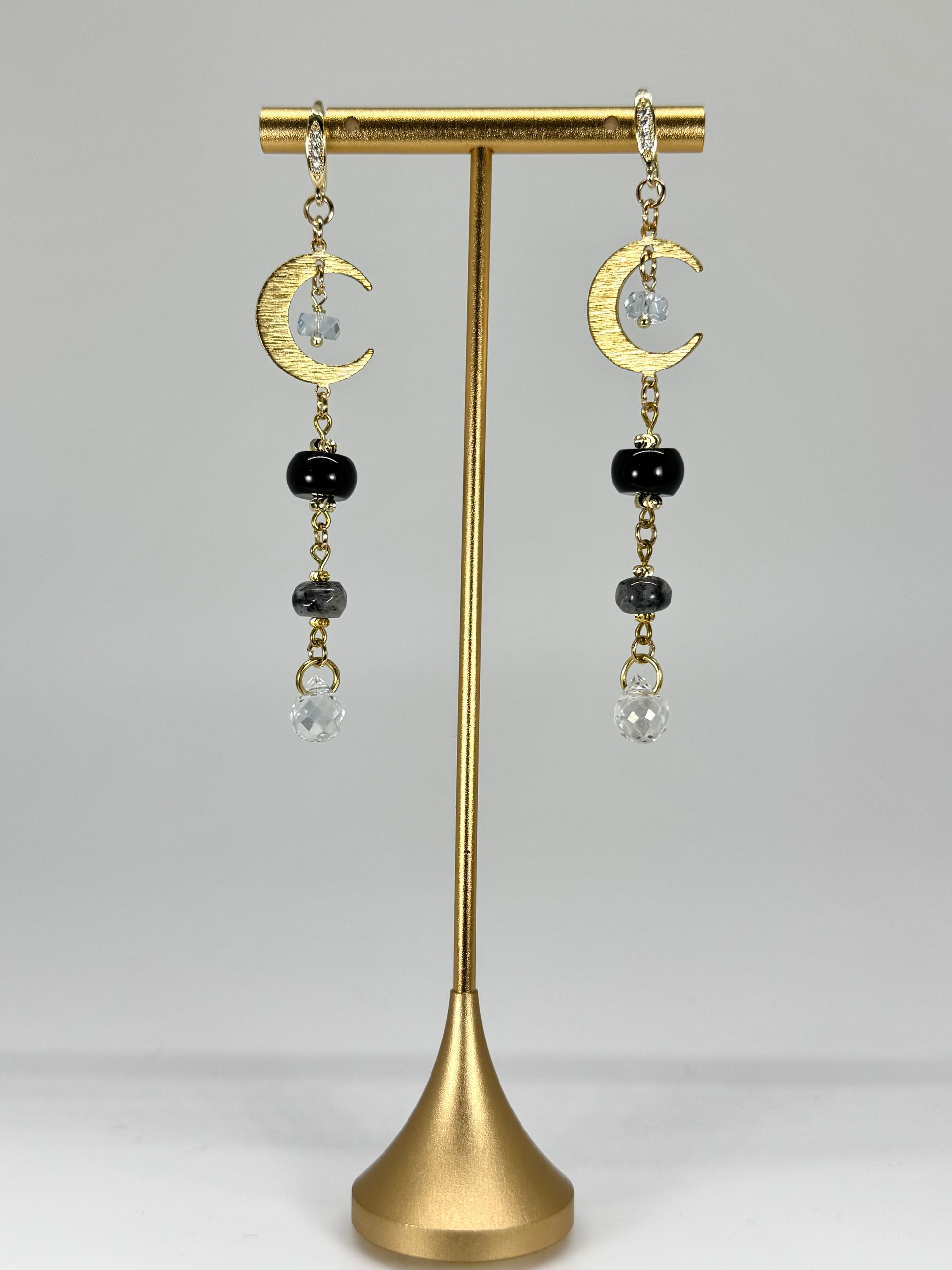 Gold earrings with crescent moon and black bead designs on a gold stand against a gray background