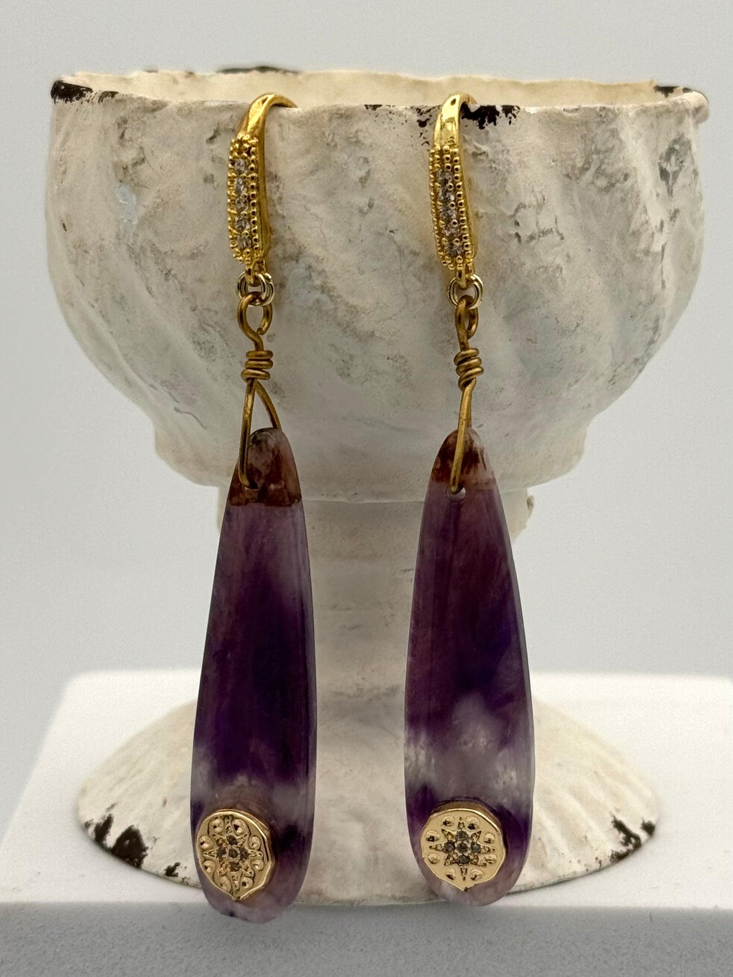 Purple teardrop earrings with gold accents on a textured white stand.