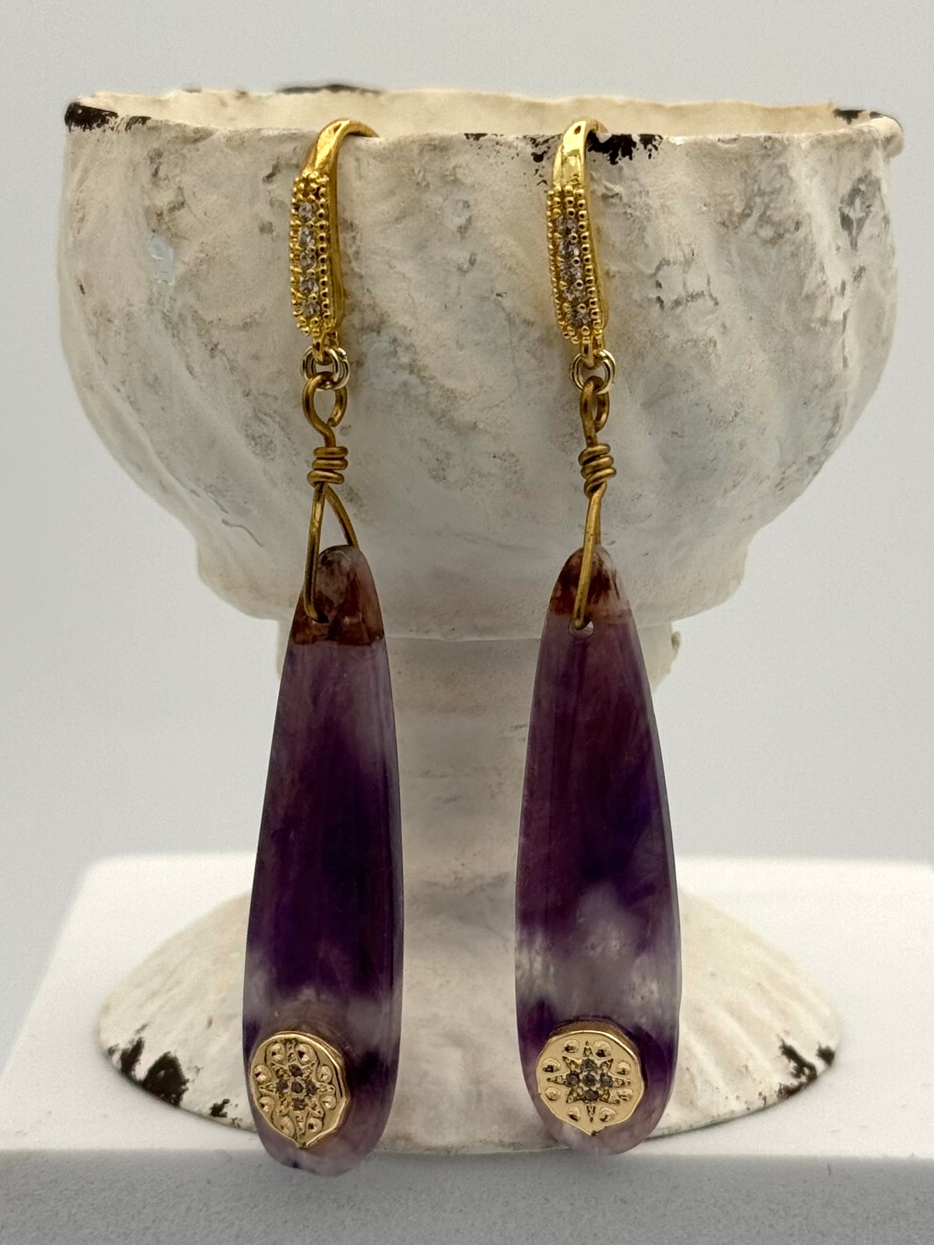 Purple teardrop earrings with gold accents on a textured white stand.