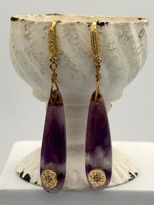 Purple teardrop earrings with gold accents on a textured white stand.
