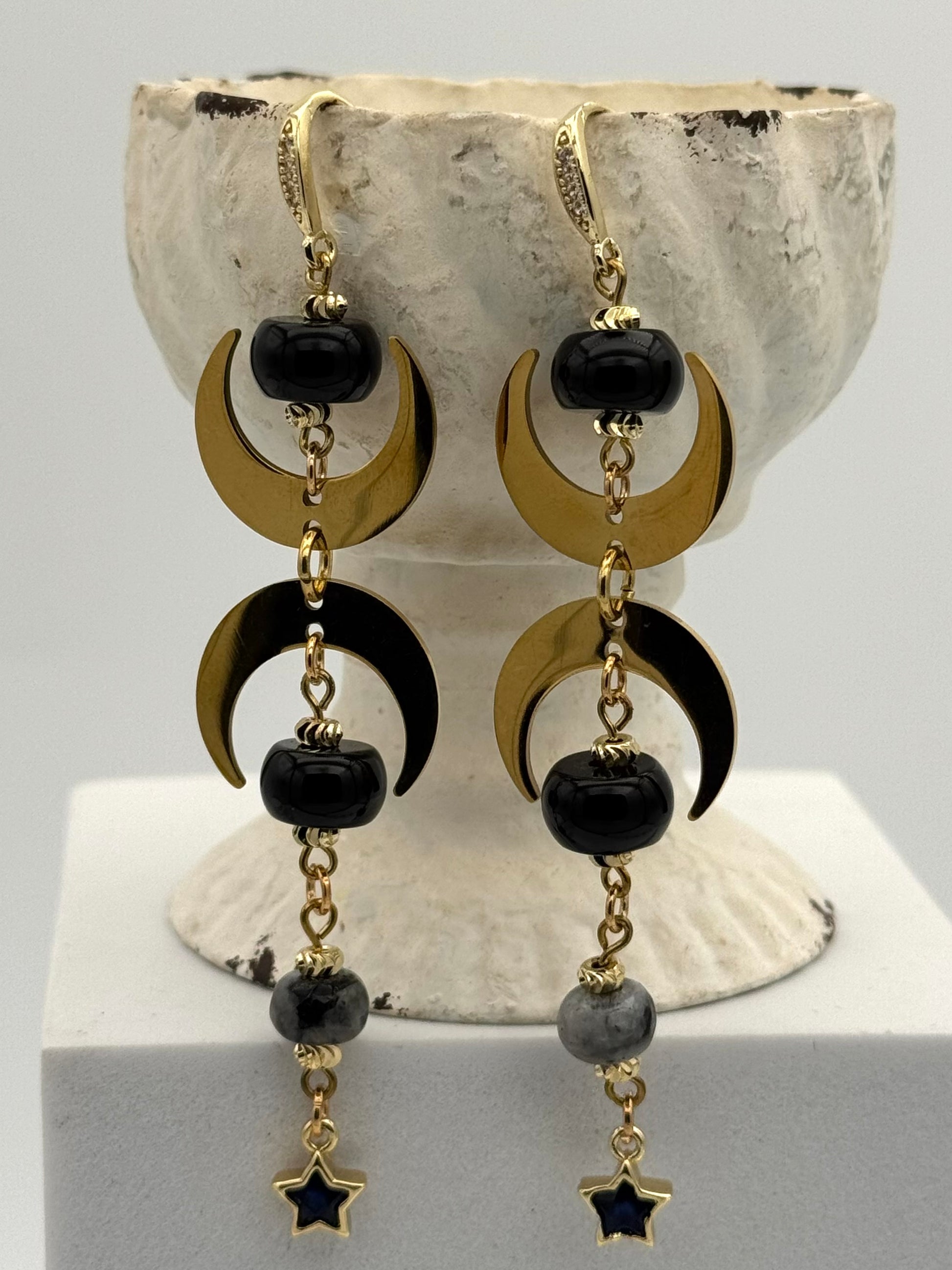Pair of earrings with black beads and gold crescent moon and star designs on a white stand.