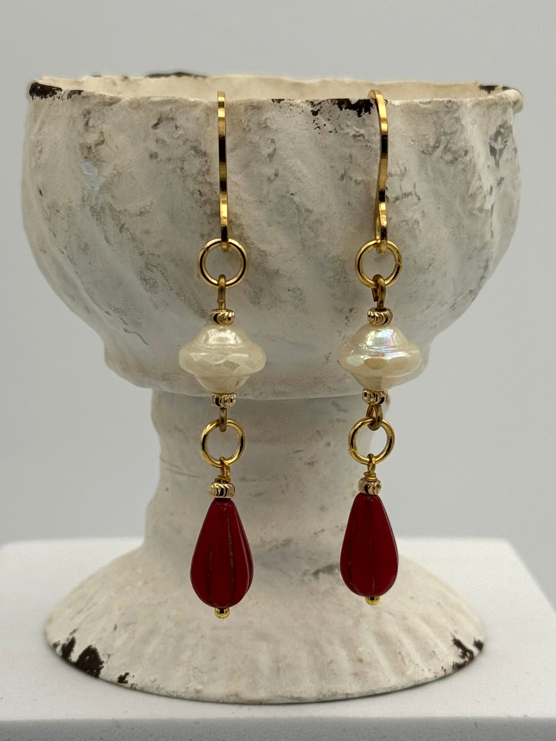 Pair of earrings with gold hooks, white beads, and red teardrop stones displayed on a textured stone surface.