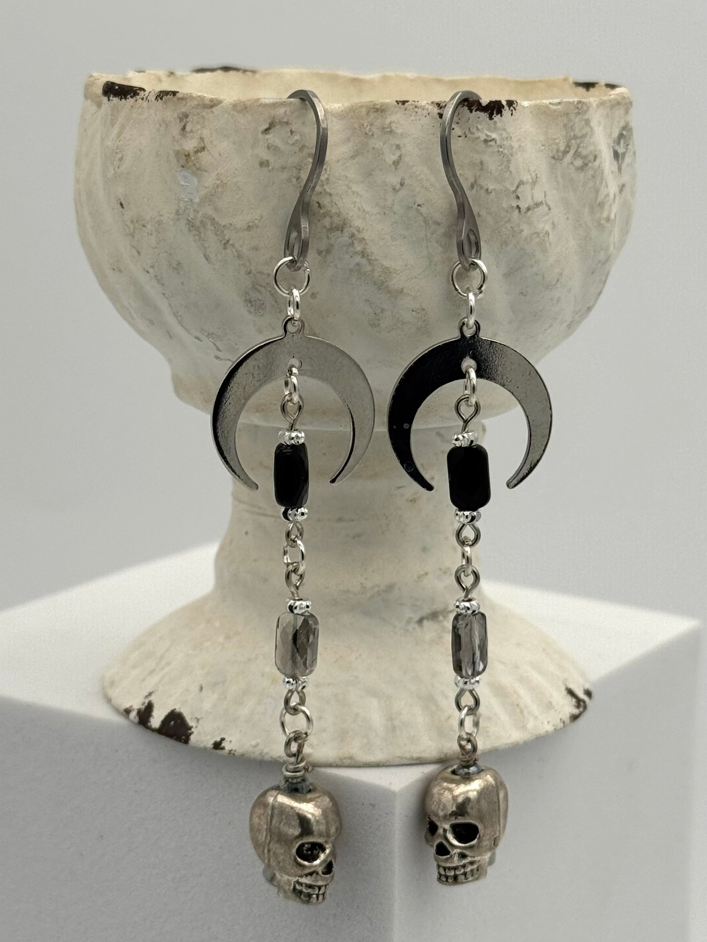 Pair of earrings with black crescent moons and skull designs on a textured white stand.