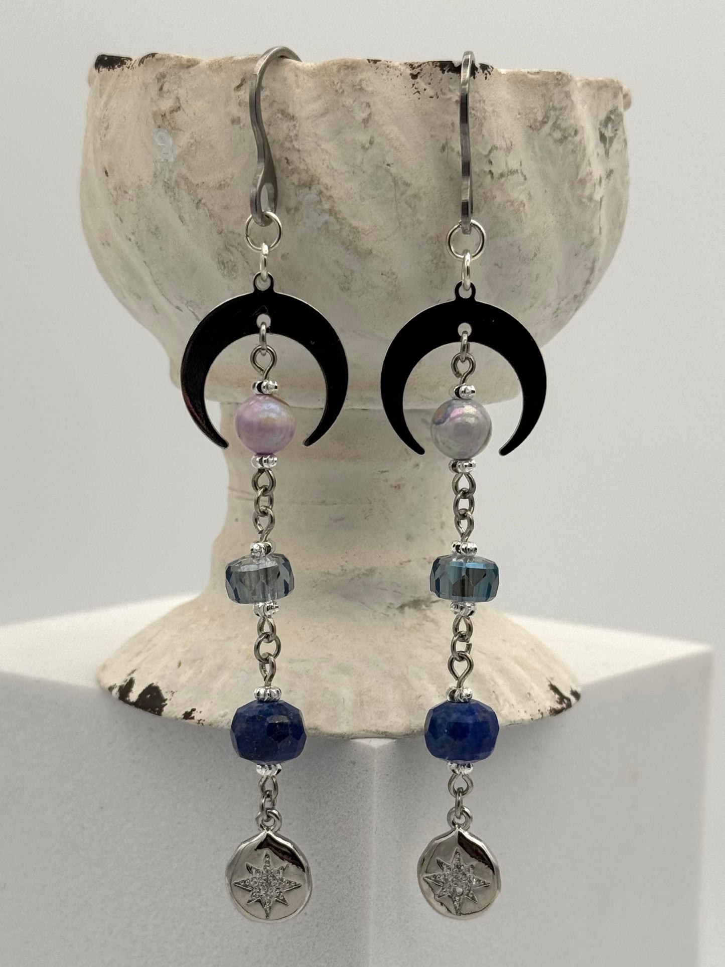 Pair of earrings with black crescent moons, blue beads, and silver pendants on a shell background.