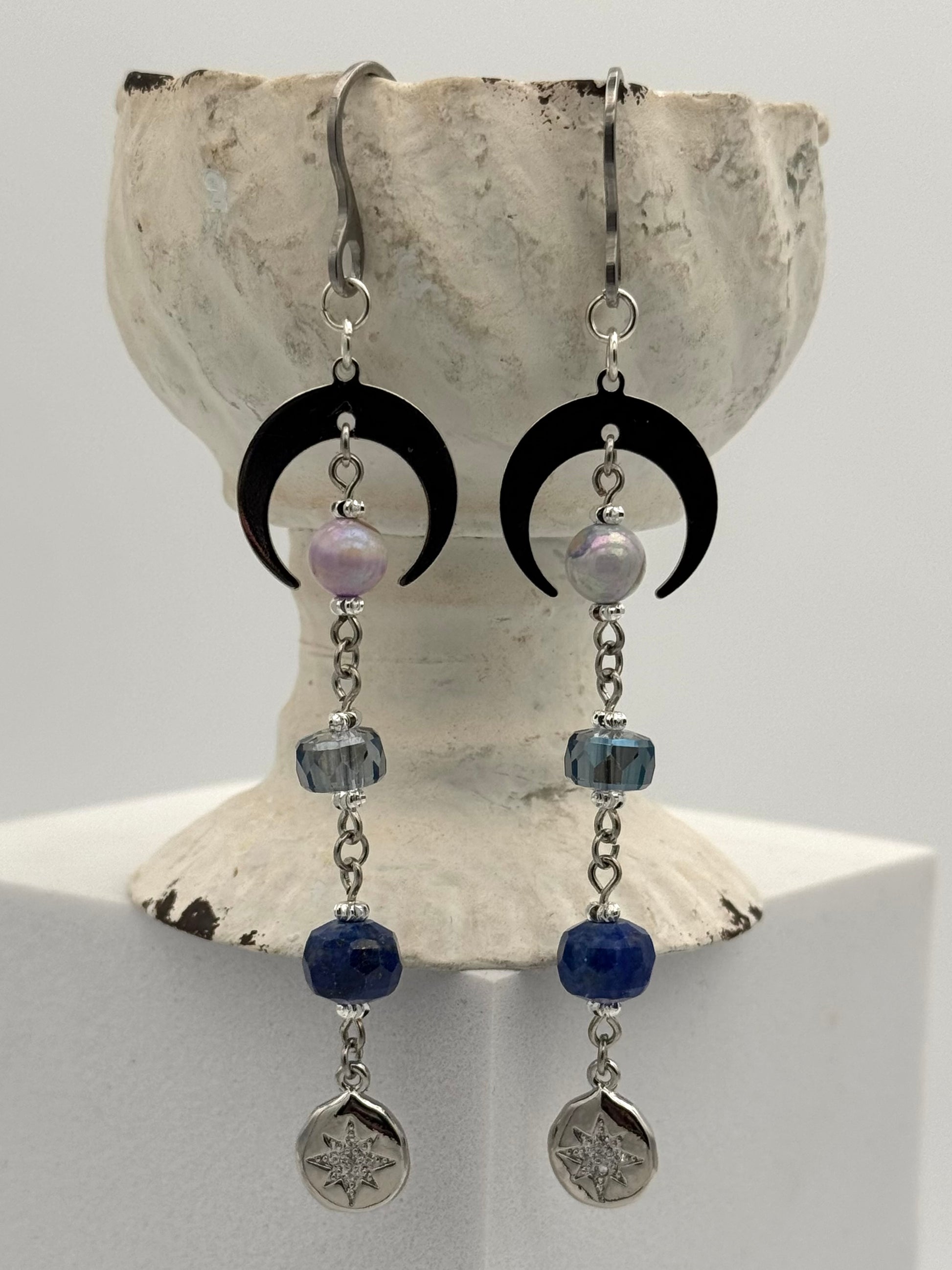 Pair of earrings with black crescent moons, blue beads, and silver pendants on a shell background.