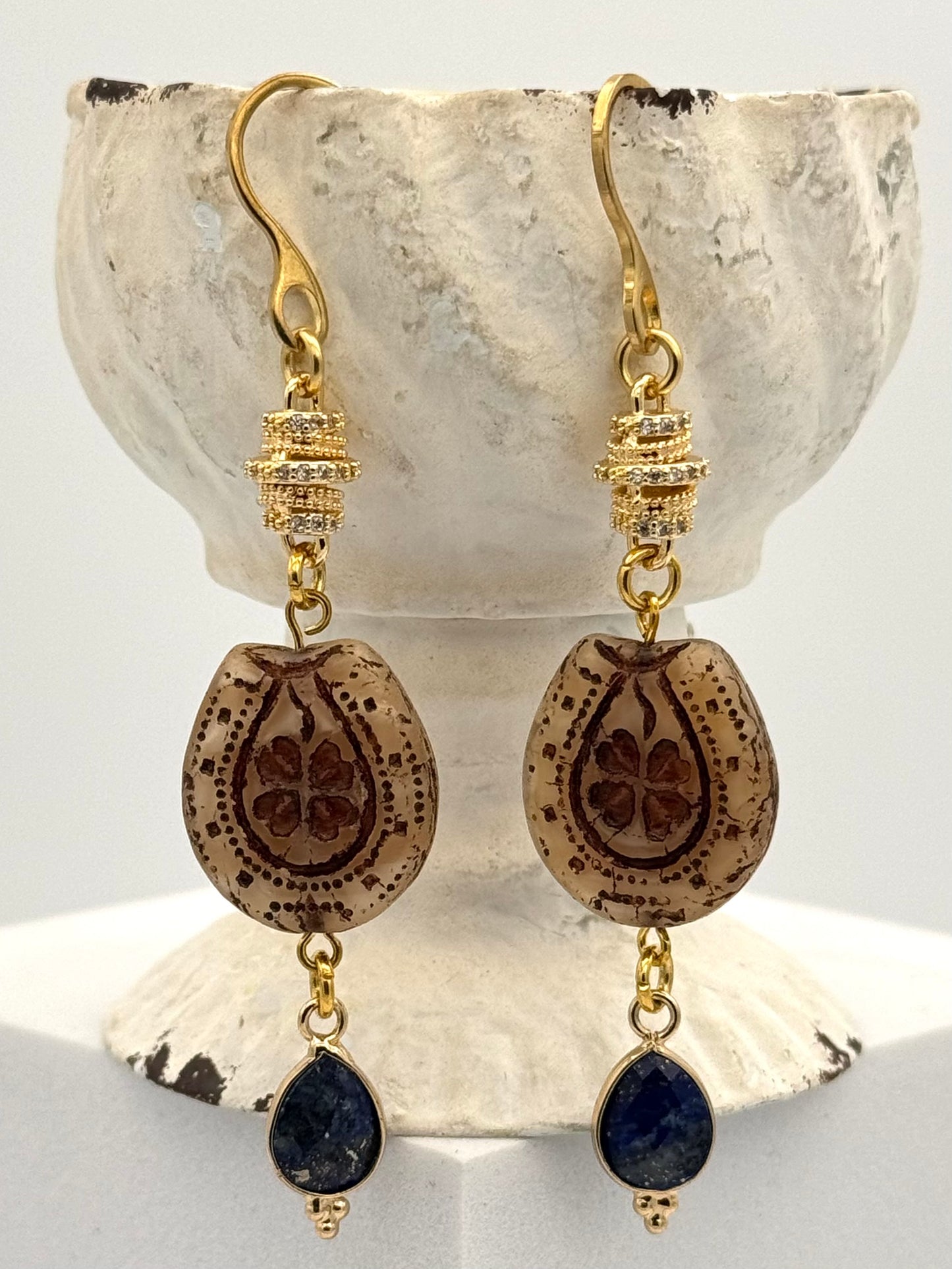 Gold earrings with brown teardrop-shaped beads and blue stones on a shell background