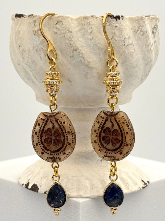 Gold earrings with brown teardrop-shaped beads and blue stones on a shell background