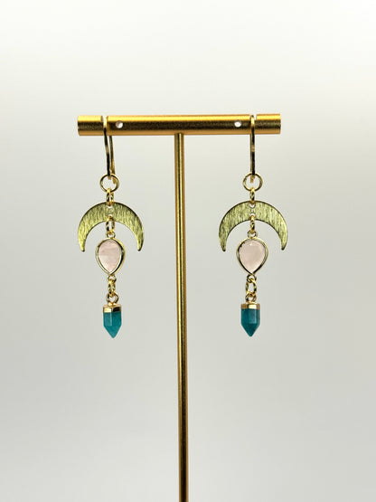 Gold earrings with moon-shaped designs and blue beads on a stand against a light background
