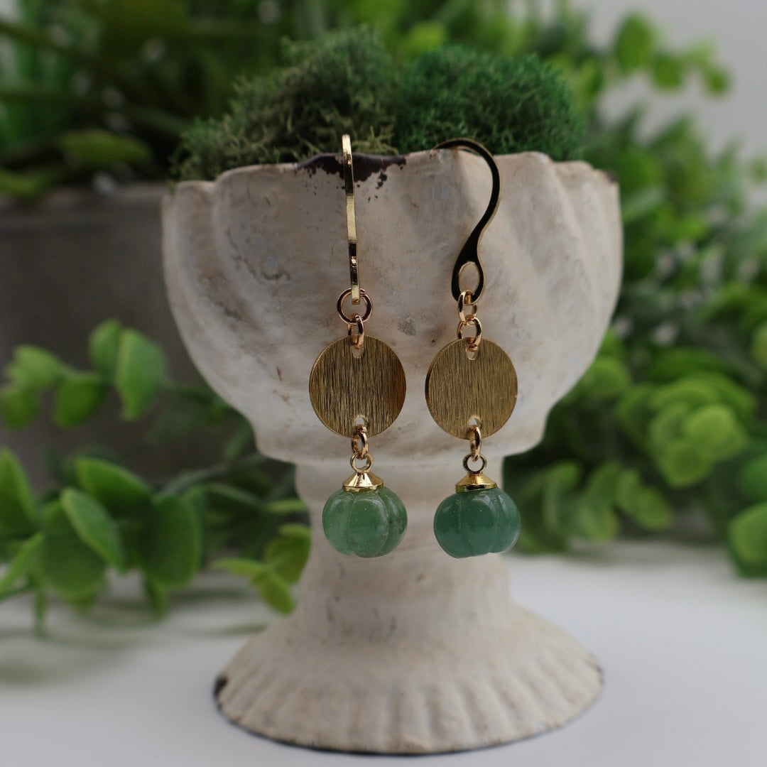 Gold earrings with green beads on a decorative stand with plants in the background