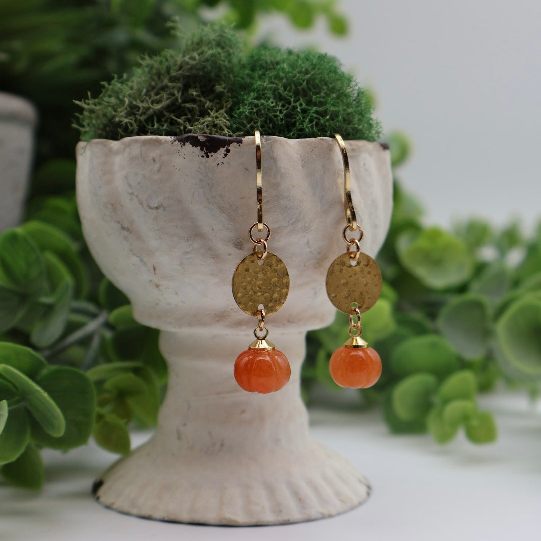 Gold and orange earrings on a white pedestal with green foliage in the background