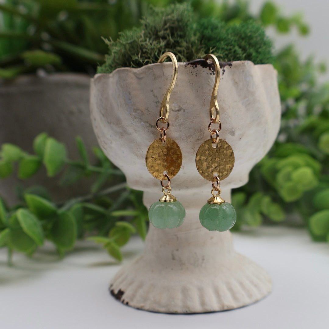 Gold earrings with green beads on a decorative stand with plants in the background