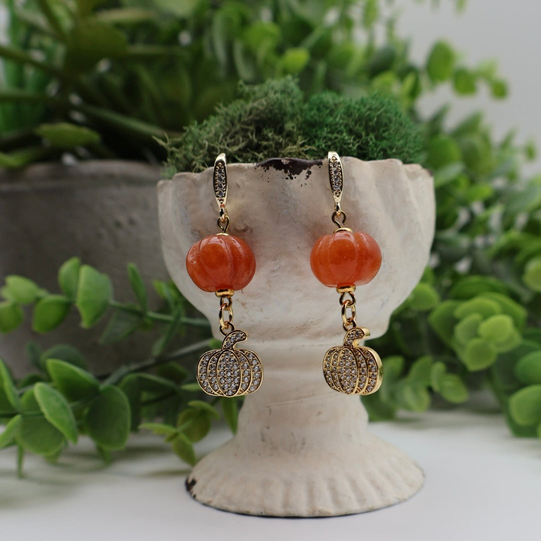 Pair of earrings with orange beads and gold accents on a decorative stand with greenery in the background.