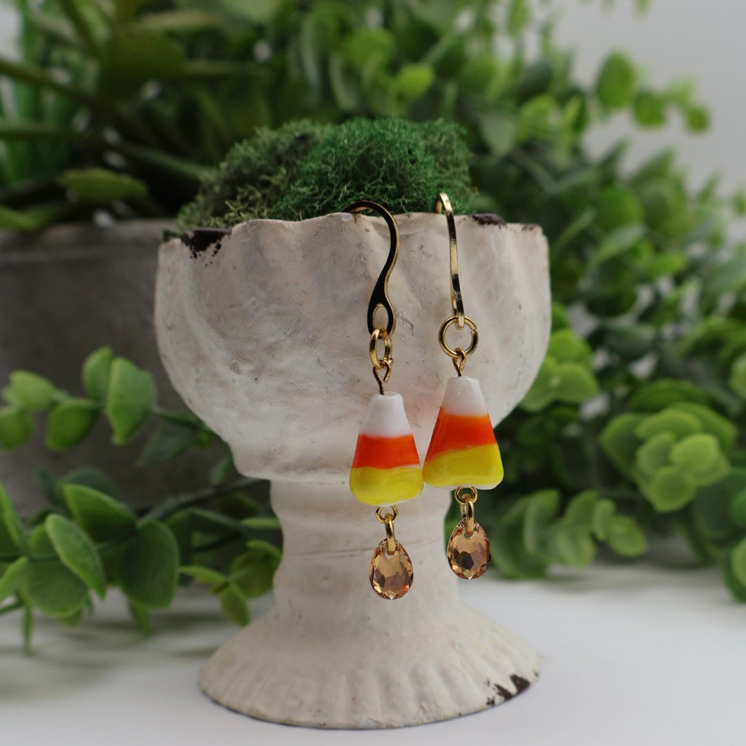 Candy corn-themed earrings on a decorative stand with greenery in the background