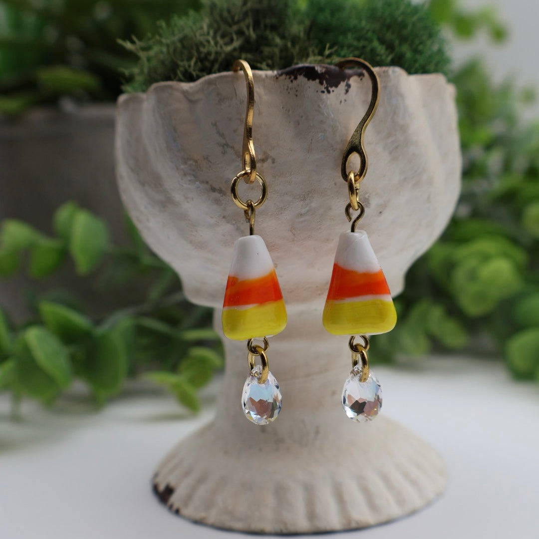 Candy corn-themed earrings with clear beads on a decorative stand with greenery in the background