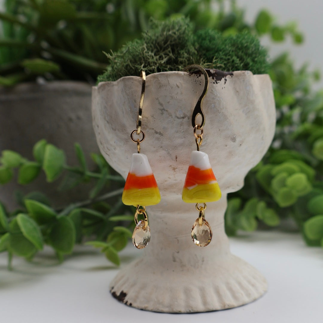 Candy corn-shaped earrings with clear teardrop beads on a white stand with greenery in the background