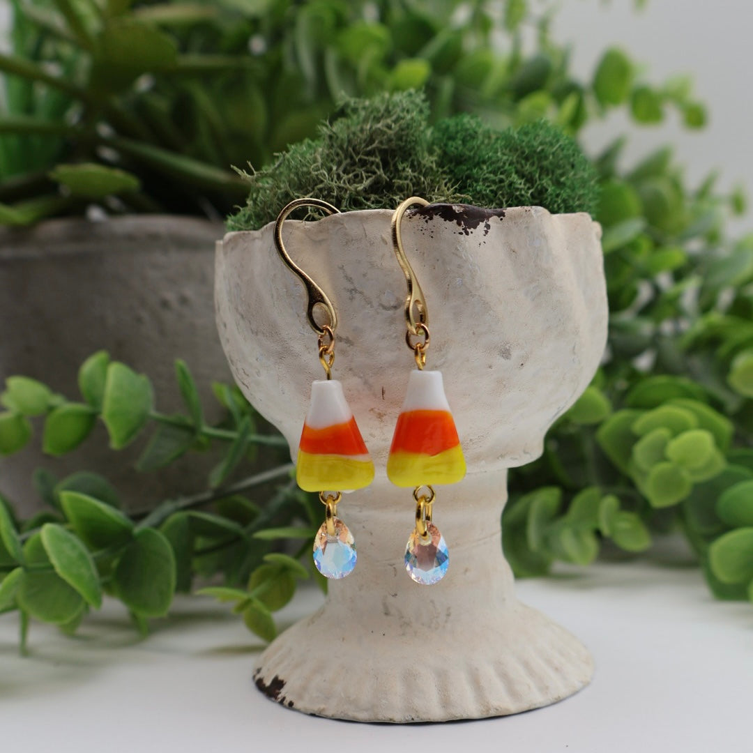 Candy corn-themed earrings with gold hooks on a decorative stand with greenery in the background.