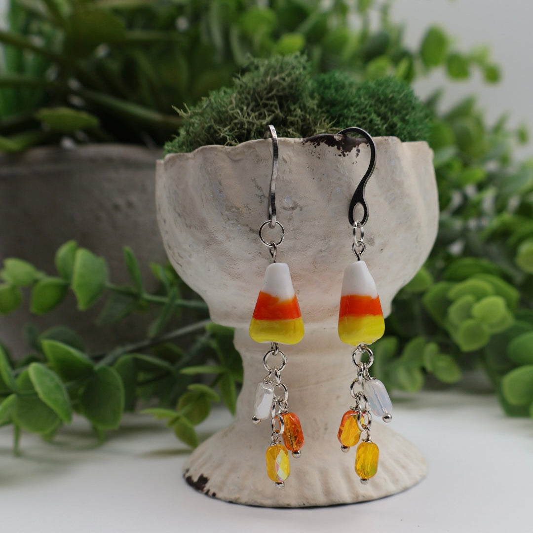 Candy corn-themed earrings on a decorative stand with greenery in the background