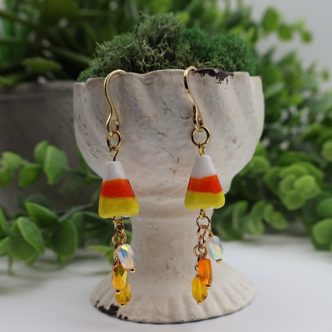 Candy corn-themed earrings on a decorative stand with greenery in the background