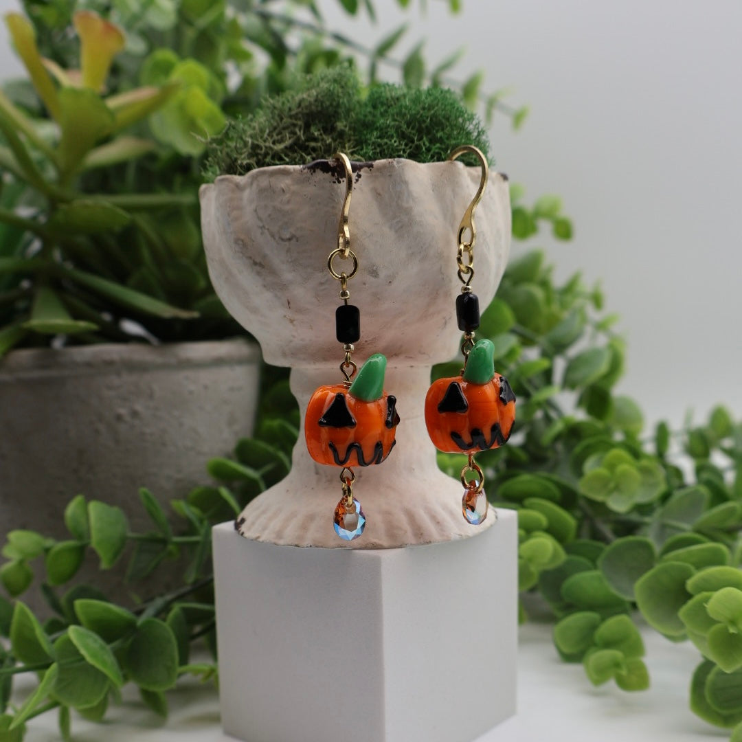 Pumpkin-shaped earrings on a decorative stand with green plants in the background