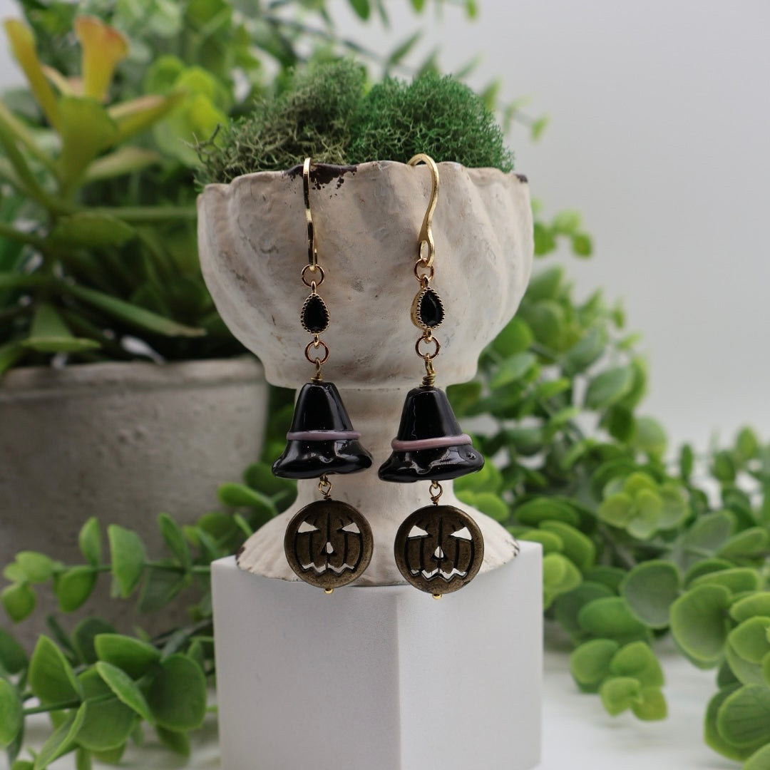 Pair of black and gold earrings on a white stand with green plants in the background