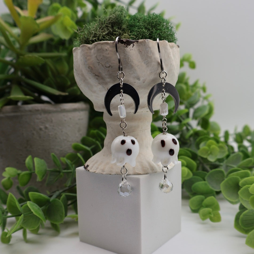 Pair of ghost-shaped earrings with crescent moon design on a white stand with green plants in the background.