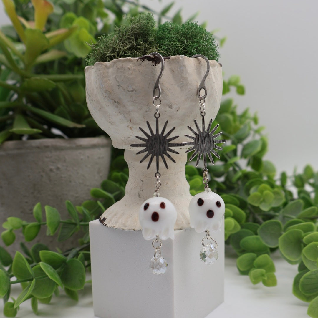 Pair of ghost-shaped earrings on a decorative stand with green plants in the background
