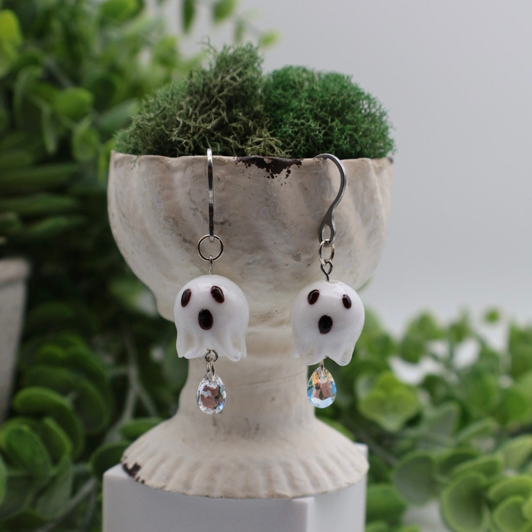 Ghost-shaped earrings with crystal drops on a decorative stand with greenery in the background