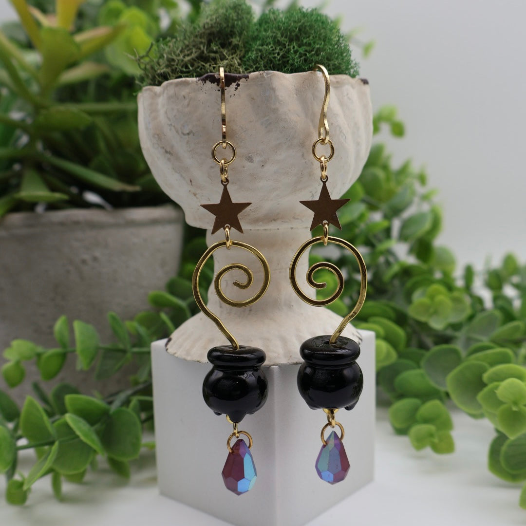 Pair of earrings with star and spiral design on a decorative stand with green plants in the background