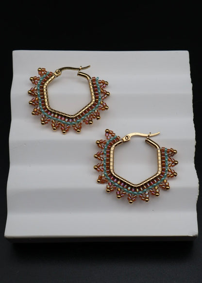 Pair of gold hoop earrings with intricate designs on a white stand against a black background