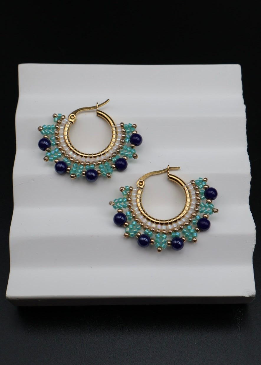 Gold hoop earrings with blue and green beads on a white stand.
